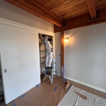 Painter in white work clothes on a ladder paints a closet in a room with wooden beams; project by Pearl Painters, Portland.