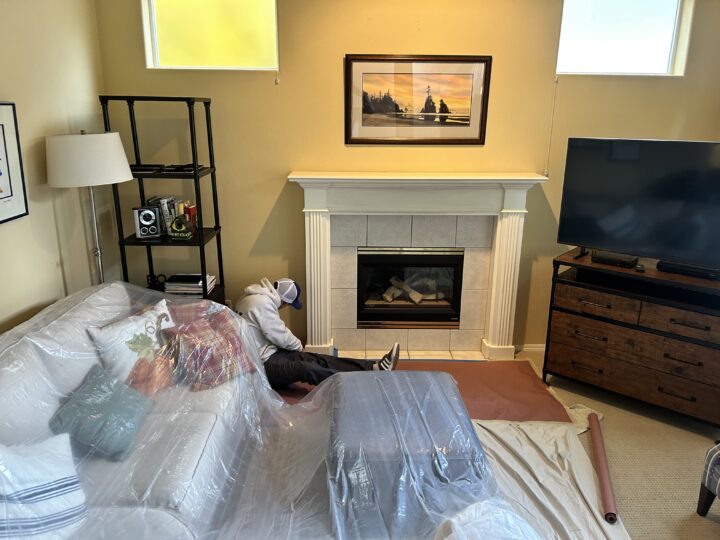 Person sitting by a fireplace in a living room, laying brown paper on the floor. The room has a covered couch, a TV, a bookshelf, and a painting above the fireplace.