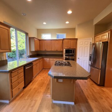 Spacious kitchen with wooden cabinetry, granite countertops, an island with a stove, stainless steel appliances, and hardwood floors. Large windows provide natural light.