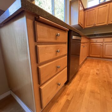 Wooden kitchen cabinets and drawers with a stainless steel dishwasher, light wood flooring, and a view of trees through the window.