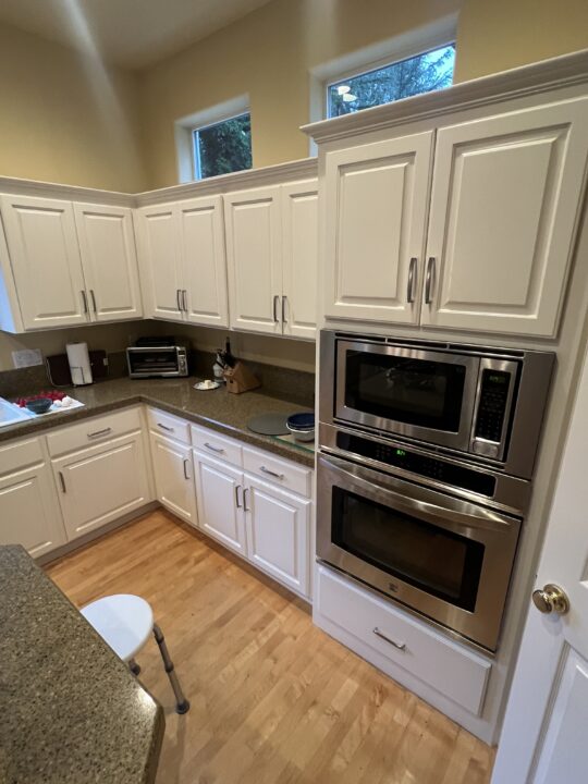 A kitchen with white cabinets, a double wall oven, granite countertops, small appliances, and a door partially visible on the right.