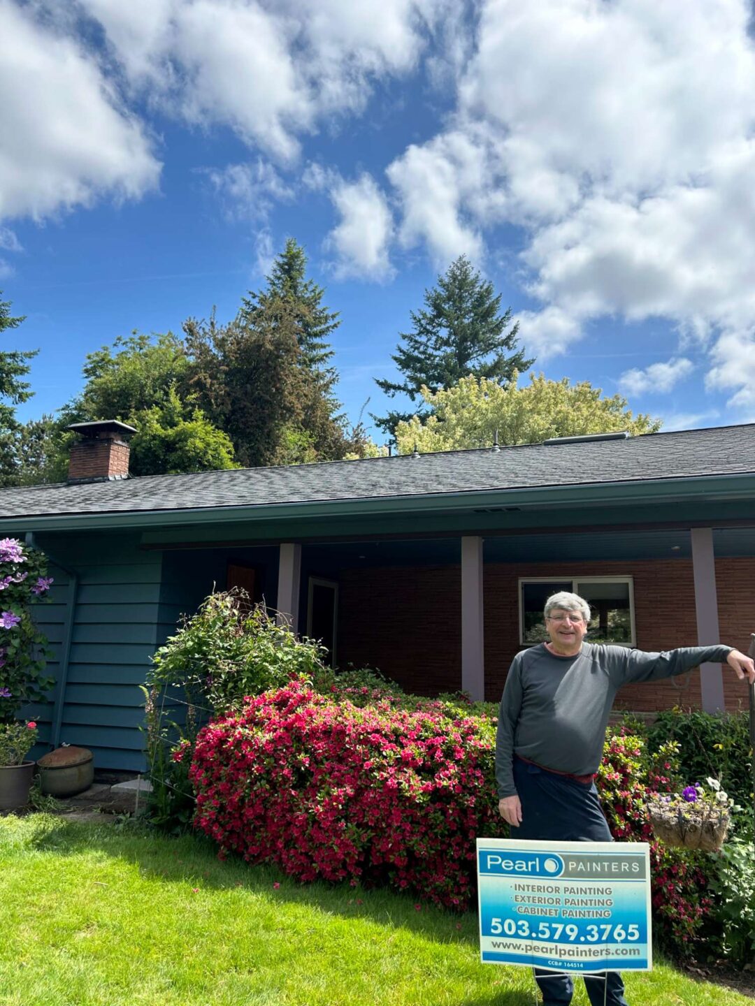 A painter stands in front of a house with blooming bushes in Portland, next to a Pearl Painters sign advertising expert interior and exterior painting services by Persichetti.