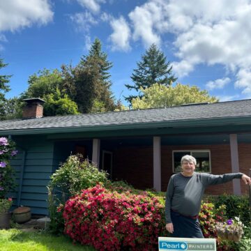 A painter stands in front of a house with blooming bushes in Portland, next to a Pearl Painters sign advertising expert interior and exterior painting services by Persichetti.