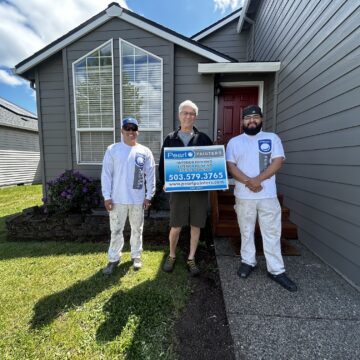 Three painters in uniform stand in front of a gray house in Horn, holding a "Peralt Painters" sign. They are ready for an exterior painting project on a sunny May 2025 day. (Not affiliated with Pearl Painters of Portland.).
