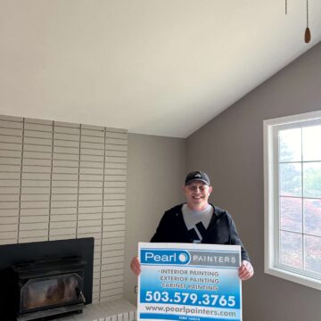 A man stands in a freshly painted Portland living room holding a Pearl Painters sign advertising interior, exterior, and cabinet painting services by painter Oneal, including phone number and website.