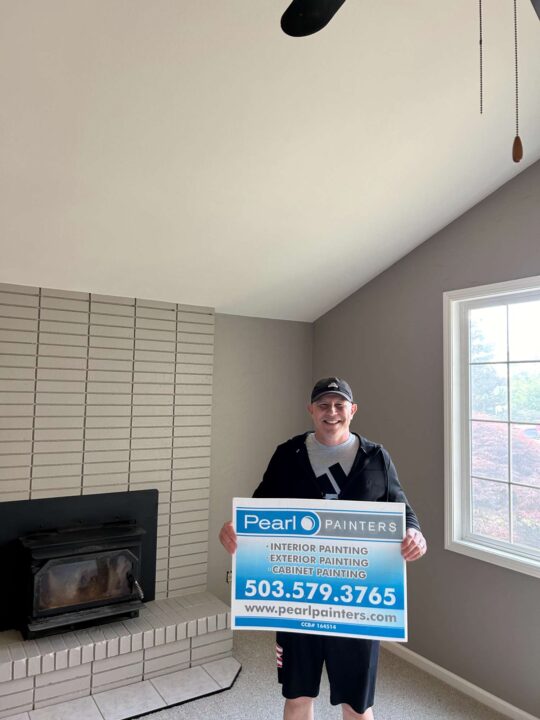 A man stands in a freshly painted Portland living room holding a Pearl Painters sign advertising interior, exterior, and cabinet painting services by painter Oneal, including phone number and website.