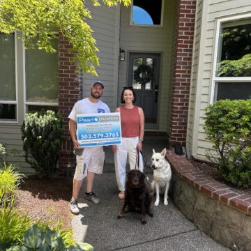 Two people stand in front of a house in Portland, holding a "Pearl Painters" sign with two dogs. The painting work was completed by painter Mackey-Hinzdel and Pearl Painters, June 2025.
