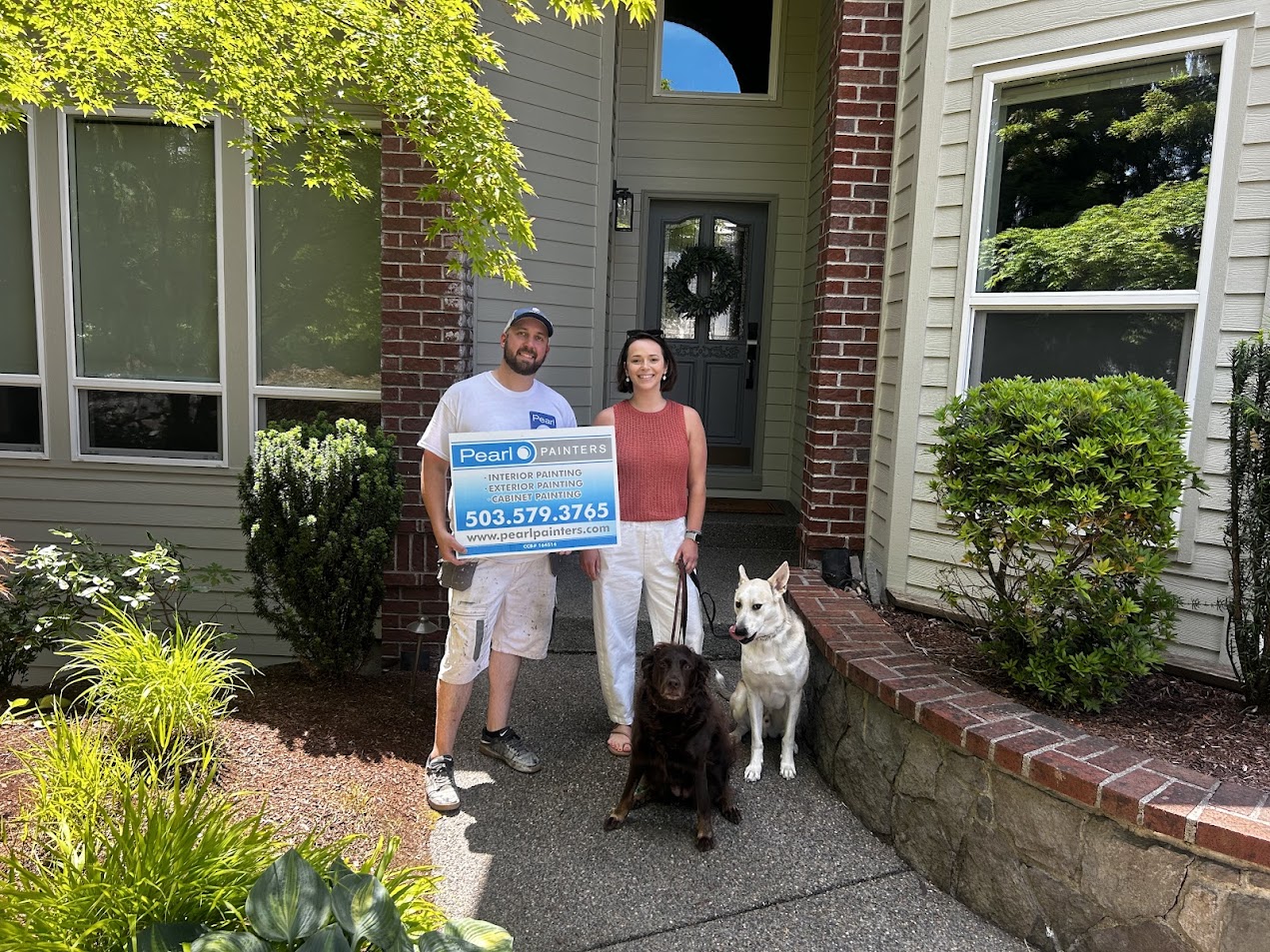 Two people stand in front of a house in Portland, holding a "Pearl Painters" sign with two dogs. The painting work was completed by painter Mackey-Hinzdel and Pearl Painters, June 2025.