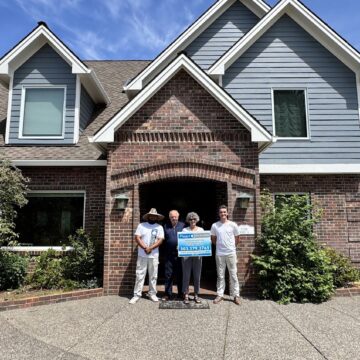 Four people stand in front of a brick house in Portland; two hold a sign with "Anderson ext," phone numbers, and other text. Photo taken June 2025, possibly for Pearl Painters or painting services.
