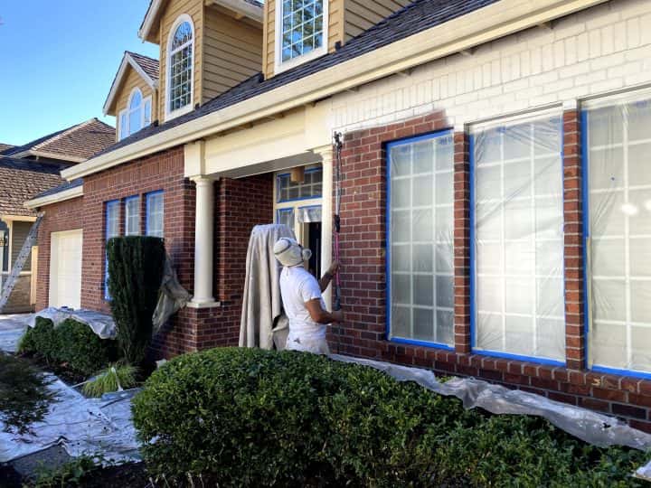 A man painting the exterior of a house with brick siding.