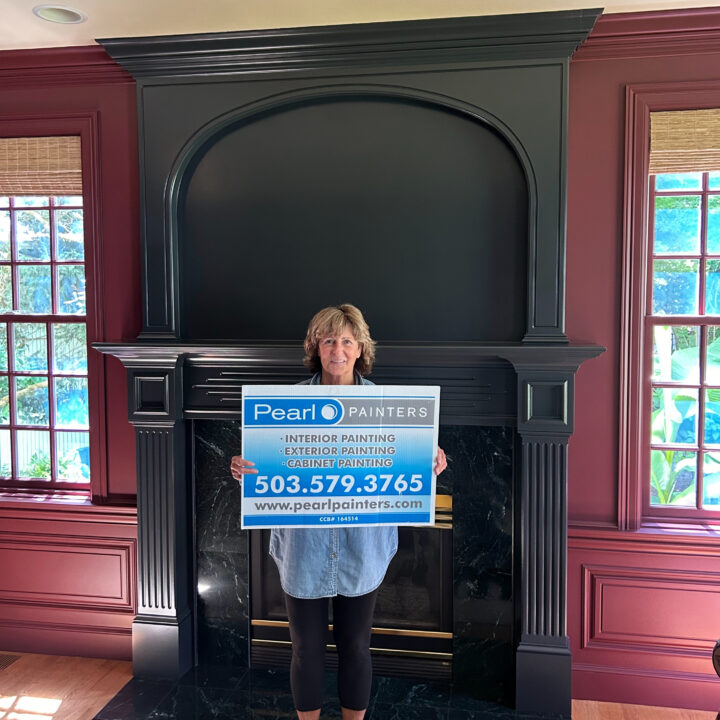Woman holds Pearl Painters sign for painting services in front of a dark fireplace; Portland painter info and phone number shown.