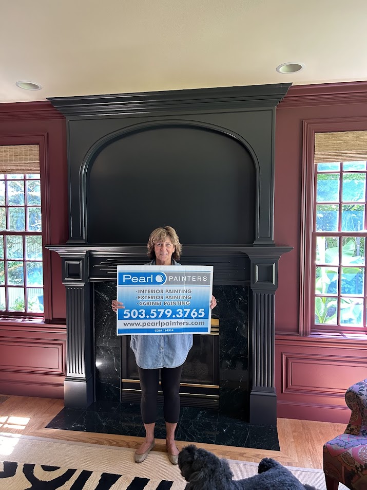 Woman holding Pearl Painters sign for interior and exterior painting services in Purcell stands by black fireplace.