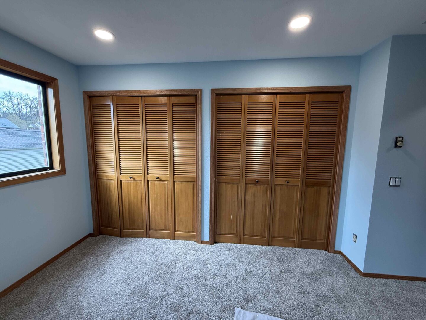 Pearl Painters painted a bright interior room with light blue walls, beige carpet, two wooden louvered closets, a window on the left, and recessed ceiling lights for the Yates residence in Portland in April 2025.