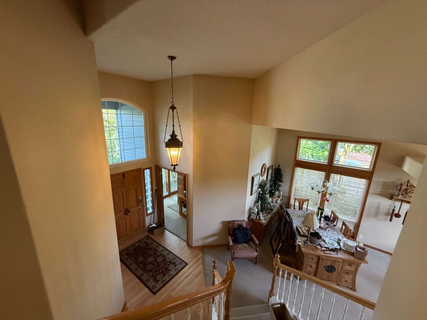 Painting of a Mann interior foyer in Portland by Pearl Painters, showing a staircase landing, wooden front door, hanging light fixture, dining area with large windows and wooden furniture in April 2025.