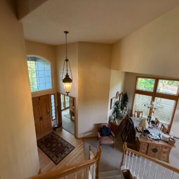 Painting of a Mann interior foyer in Portland by Pearl Painters, showing a staircase landing, wooden front door, hanging light fixture, dining area with large windows and wooden furniture in April 2025.