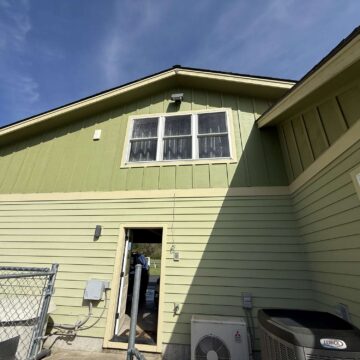 Photo of a light green house exterior with two upper windows, HVAC units, and a fence in Portland, taken by painter McCaffery in April 2025. Pearl Painters project.