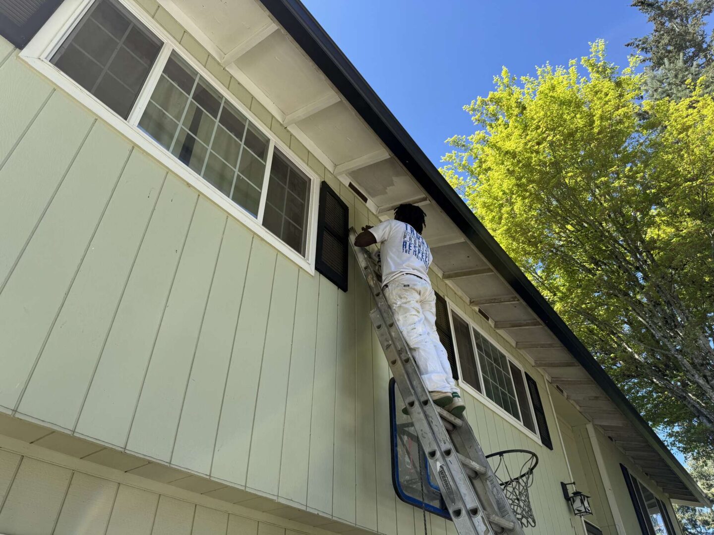 A painter in white work clothes stands on a ladder painting the exterior eaves of a light green house with large windows in Portland, likely as part of an April 2025 Pearl Painters project.