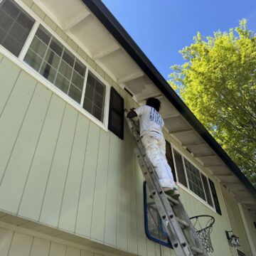 A painter in white work clothes stands on a ladder painting the exterior eaves of a light green house with large windows in Portland, likely as part of an April 2025 Pearl Painters project.