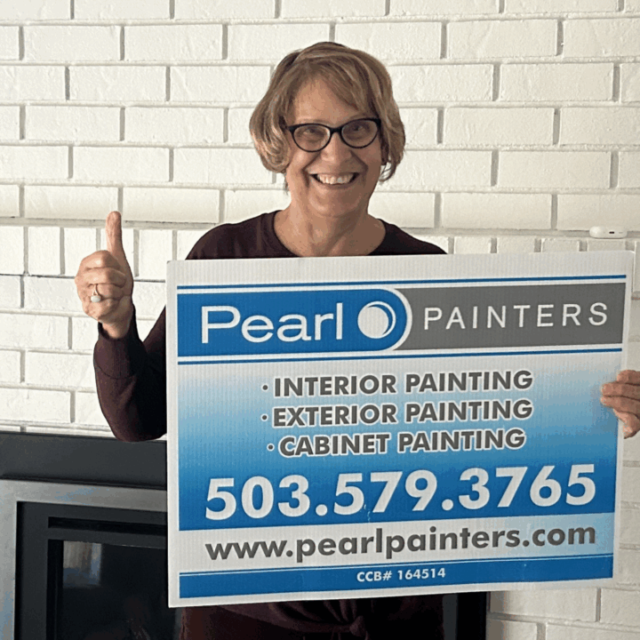 Woman holds Pearl Painters sign in front of white brick wall, advertising Portland painter services and contact info.
