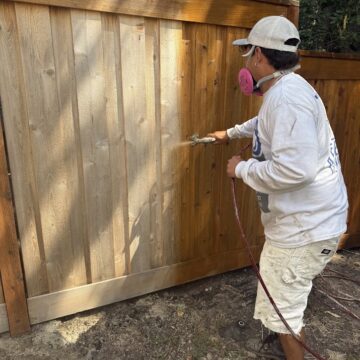 Painter Smith with protective gear stains a wooden fence using a spray gun for Pearl Painters, Portland, July 2025.