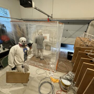 Two workers in protective gear spray painting cabinets in a plastic-enclosed setup inside a garage, with tools and equipment scattered around.