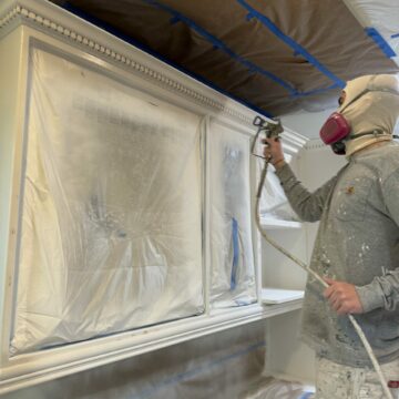 Worker in protective gear spray painting a window frame, area covered with protective plastic and tape.