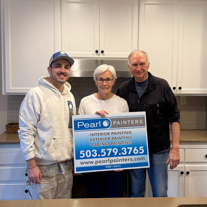 Three people in a kitchen hold a Pearl Painters sign listing painting services and contact info. Based in Portland.