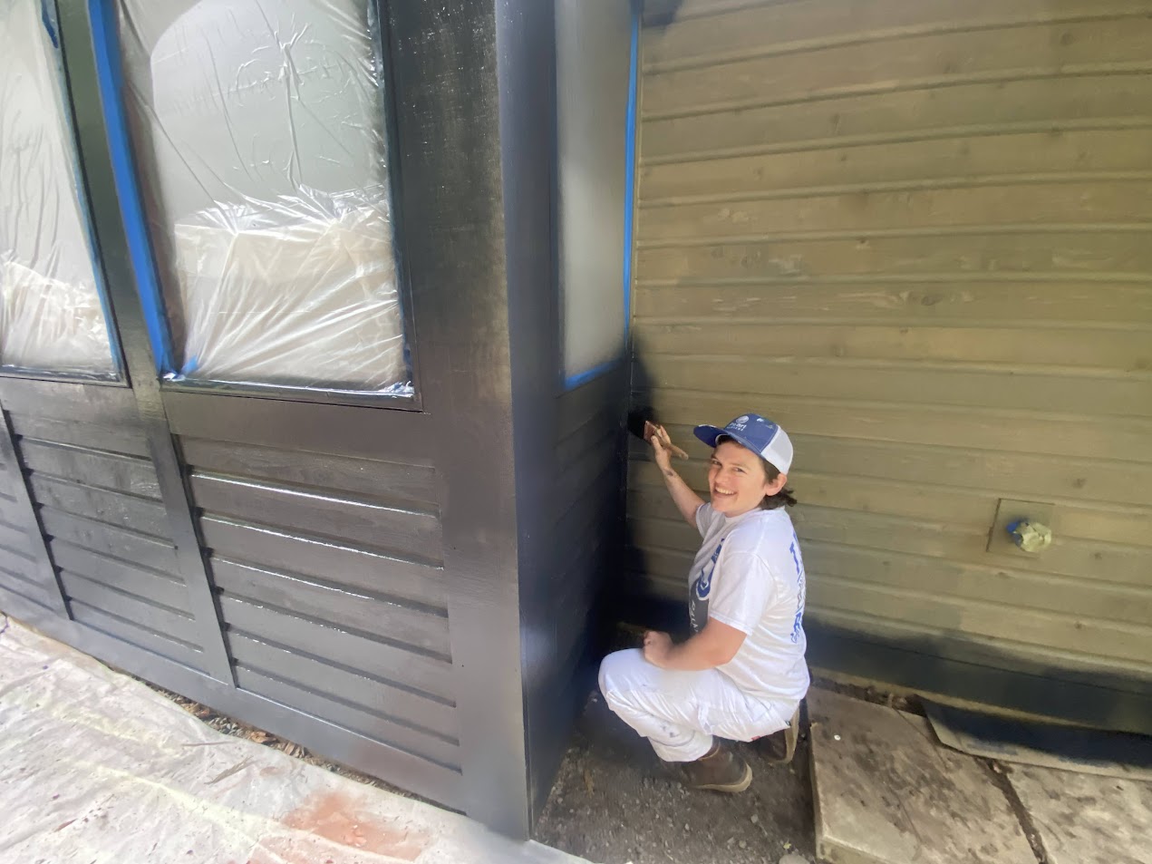 Painter in white attire paints building wall near windows taped and covered, work by Pearl Painters in Portland.