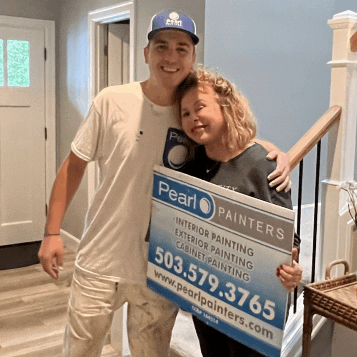 Two people indoors hold a Pearl Painters sign for painting services in Portland, with company website and phone number visible.