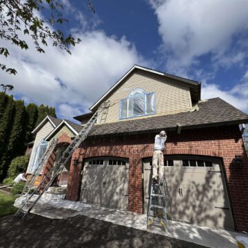 Painter Scott stands on a ladder painting exterior siding above the garage of a two-story brick house in April 2025. This work is done by Pearl Painters in Portland under partly cloudy skies.