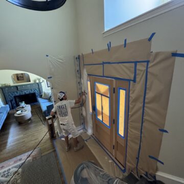 Painter in hat uses sprayer to paint front door in July 2025; walls are taped off. Project by Pearl Painters, Portland.