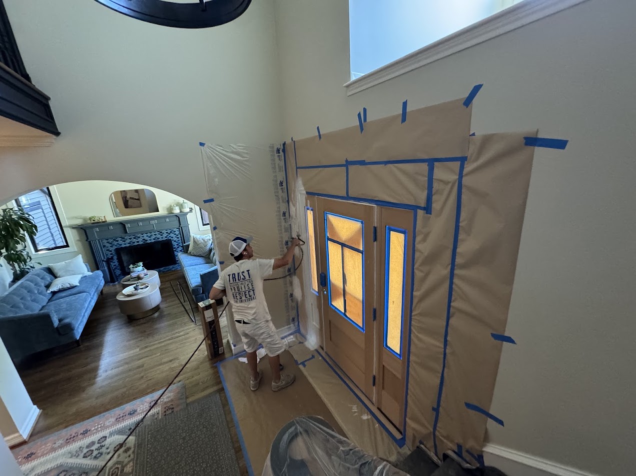 Painter in hat uses sprayer to paint front door in July 2025; walls are taped off. Project by Pearl Painters, Portland.