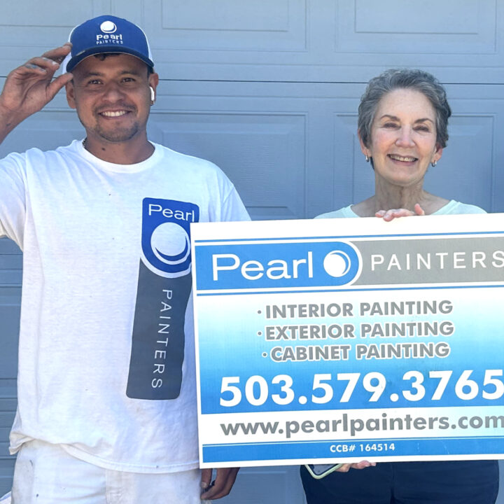 Two people stand before a gray garage; one holds a Pearl Painters sign for painting services in Portland.