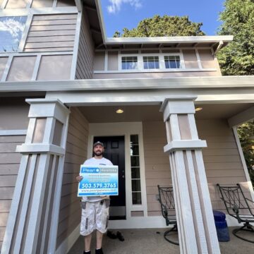 Man on beige house porch in Portland holds Pearl Painters sign about exterior painting, Anderson, and August 2025 projects.