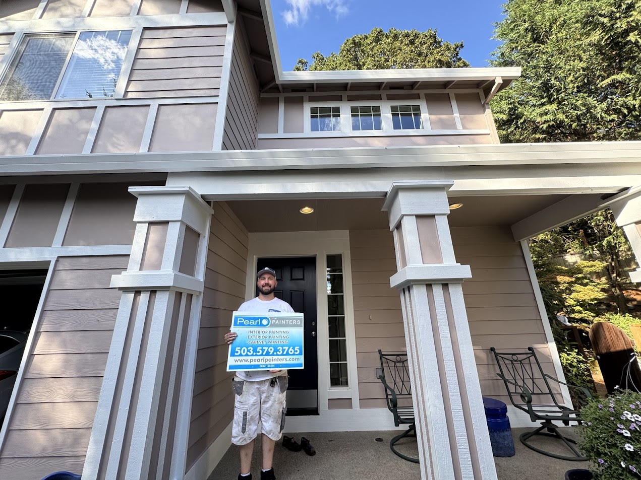 Man on beige house porch in Portland holds Pearl Painters sign about exterior painting, Anderson, and August 2025 projects.
