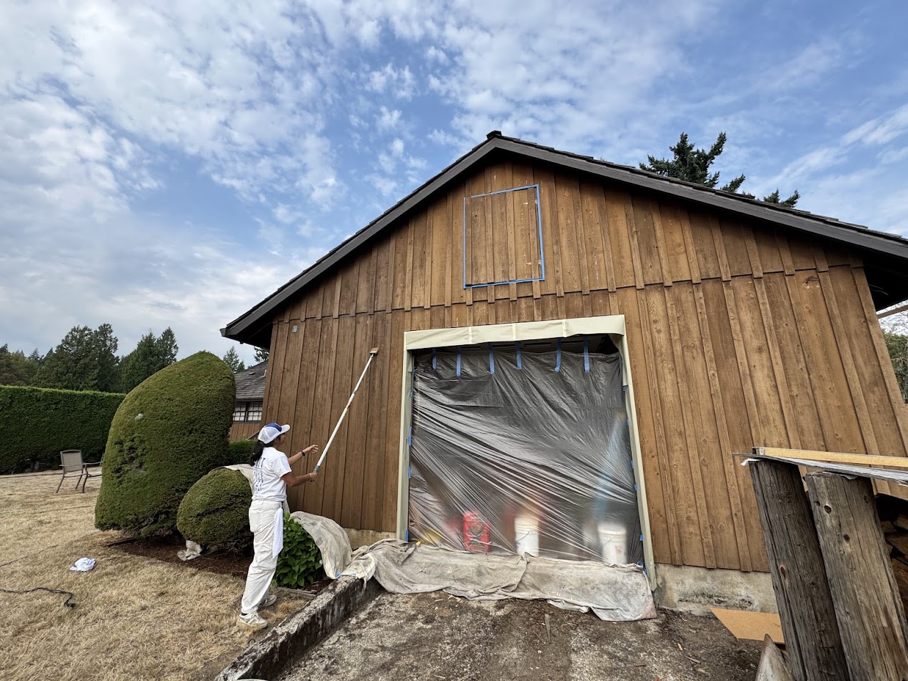 Painter in white uses roller on exterior wall; plastic covers doorway, drop cloths on ground. Pearl Painters project, Portland, Aug 2025.