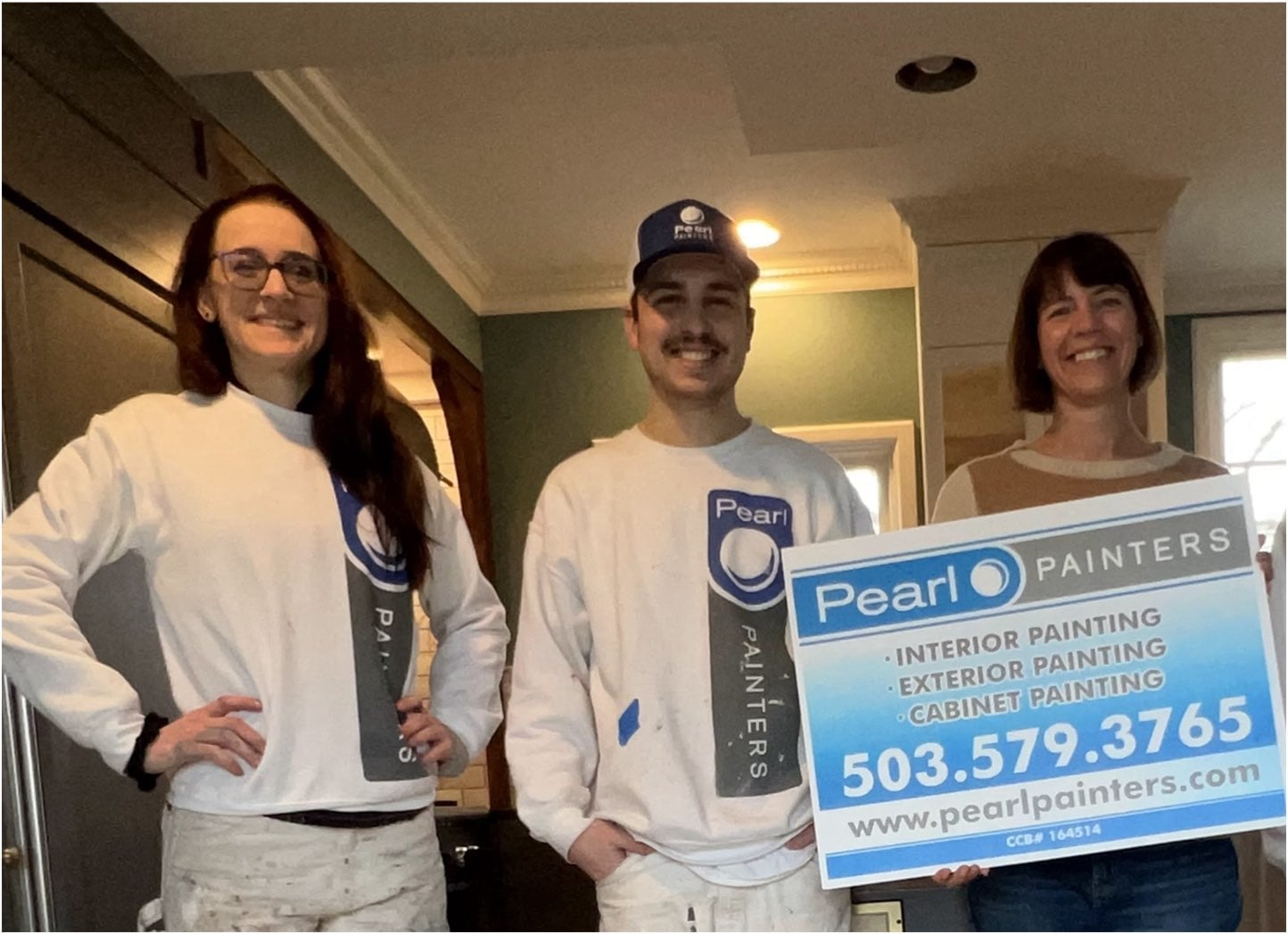 Three people stand indoors, two in painters' uniforms and one holding a sign for "Pearl Painters" with contact information and services listed.