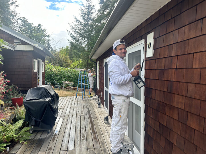 Two people are painting a house's exterior. One is on a ladder on the porch, while the other is in the background near a fence, preparing for some deck staining. Both are dressed in white work clothes.