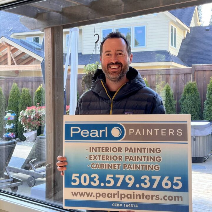 Man holds Pearl Painters sign indoors, advertising Portland painting services and contact info.