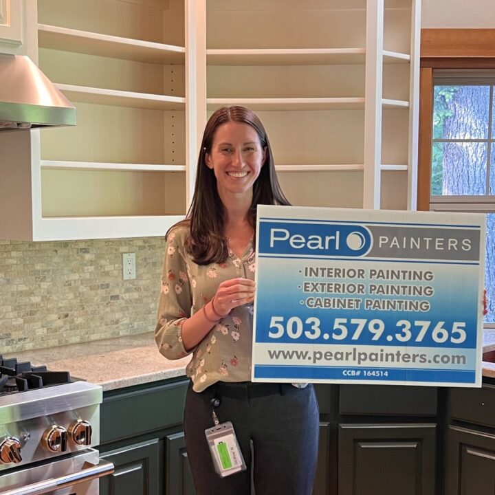 Woman in kitchen holds Pearl Painters sign for painting services in Portland, showing phone number and website.
