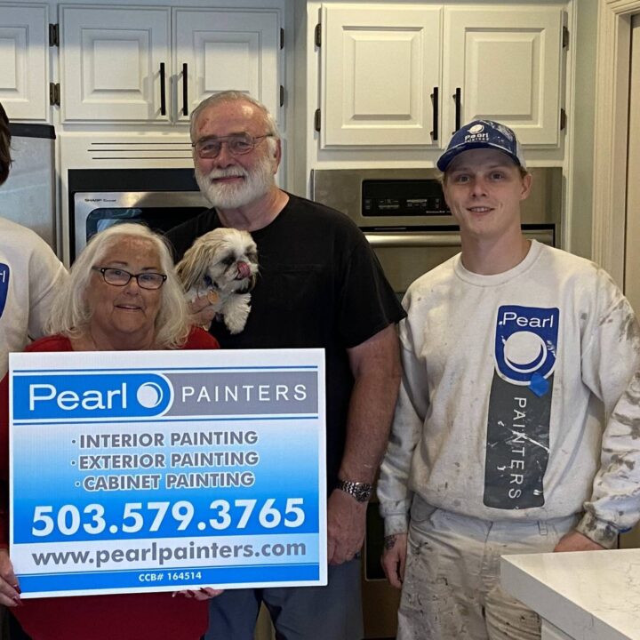 Two painters with an elderly couple and dog in a kitchen hold a Pearl Painters sign for painting services in Portland.