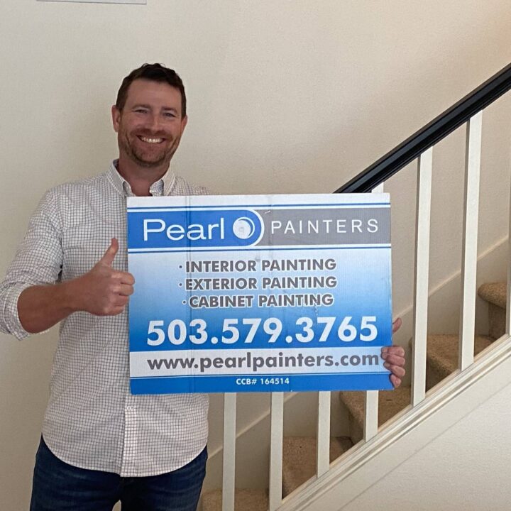 Man indoors by staircase holds Pearl Painters sign with service details, giving thumbs up. Painting company is based in Portland.