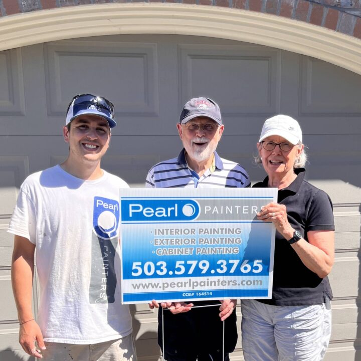 Three people in front of a garage door hold a Pearl Painters sign with service info, promoting painting in Portland.