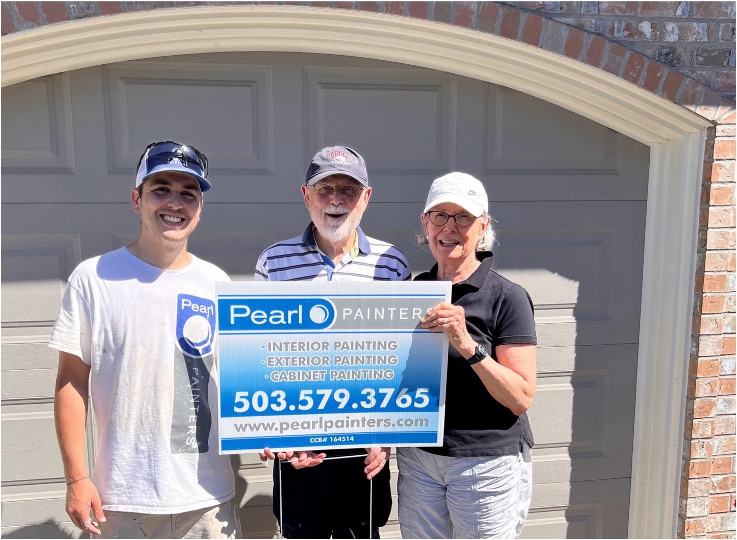 Three people stand in front of a garage door, holding a sign for "Pearl Painters" displaying services and a contact number. They are smiling and appear to be outdoors on a sunny day.