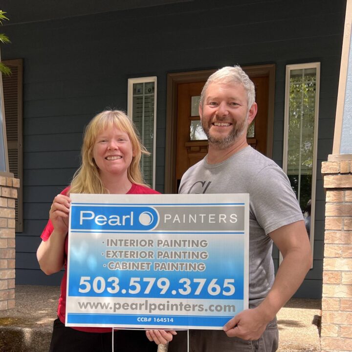 Two people in front of a house hold a Pearl Painters sign for painting services in Portland, with contact info visible.