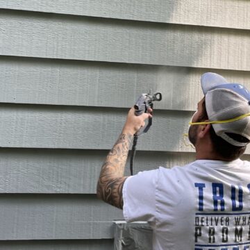 A man wearing a hat, mask, and gloves uses a paint sprayer to apply paint to horizontal siding on a building, carefully considering how many coats of paint are needed for optimal coverage.