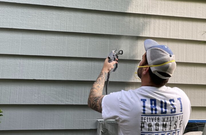 A man wearing a hat, mask, and gloves uses a paint sprayer to apply paint to horizontal siding on a building, carefully considering how many coats of paint are needed for optimal coverage.