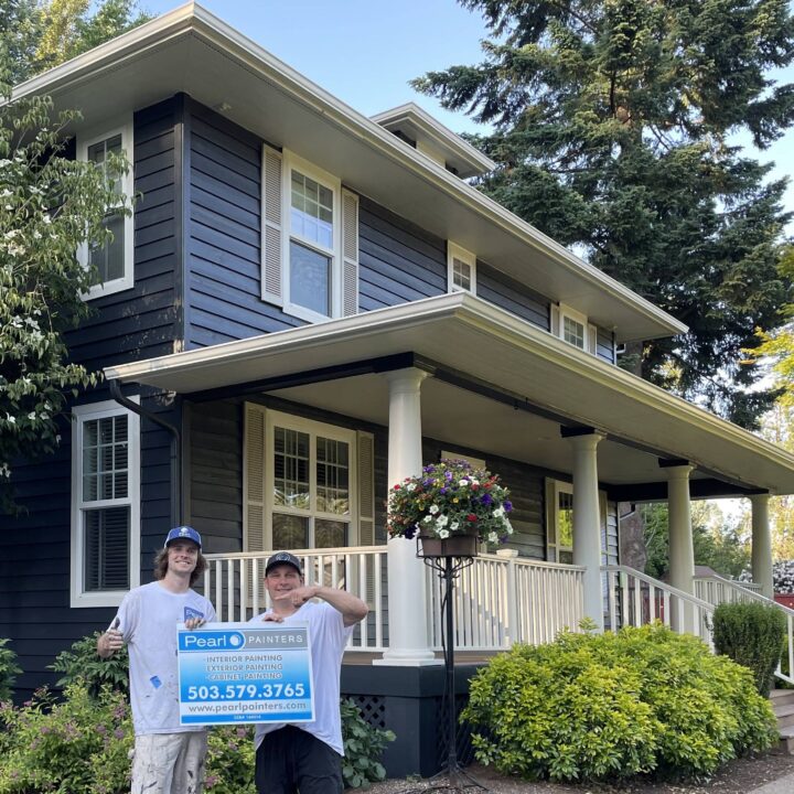 Two people with a Pearl Painters sign stand by a dark-painted house and garden, likely in Portland, on a sunny day.