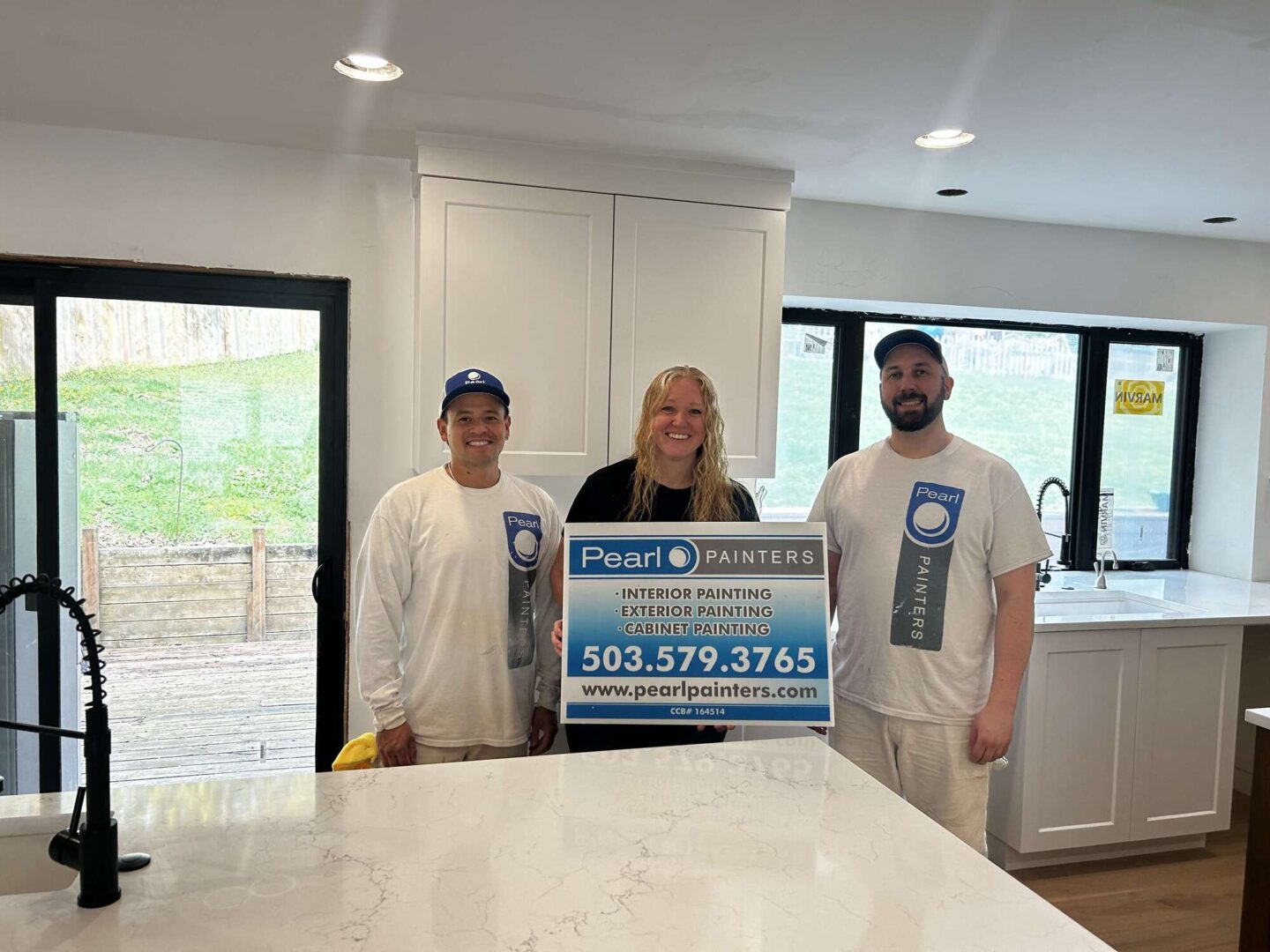 Three painters—two in white uniforms, one in black—stand in a modern kitchen holding a Pearl Painters sign with contact details and the name Evans. The location is Portland.