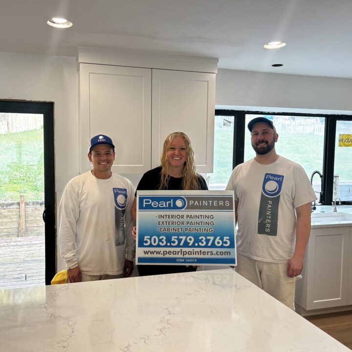 Three painters in Pearl Painters uniforms stand in a kitchen, holding a painting services sign with contact info in Portland.